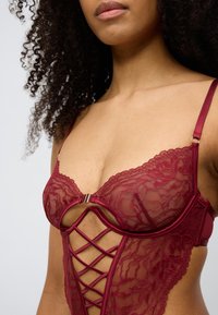 Μπορντό κορμάκι από δαντέλα με διάφανο ύφασμα, σατέν ράντες και σχέδιο σταυρωτό στο μπροστινό μέρος με χρυσή διακόσμηση.