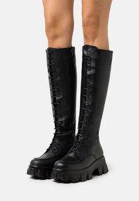 Nohy obuté v černých vysokých č lace-up botách s tlustou chunky podrážkou, na jednobarevném světlém pozadí.