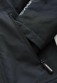 Giacca nera con tessuto testurizzato a motivo a griglia, chiusura con zip e dettagli del logo sul colletto. Il manico della zip è marchiato "Superdry."