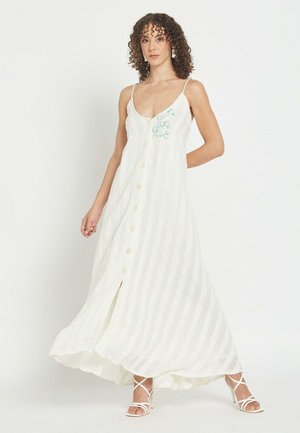 Ženska s kodrastimi lasmi, oblečena v dolgo belo obleko brez rokavov z gumbi spredaj in belimi sandali na visoko peto z naramnicami.