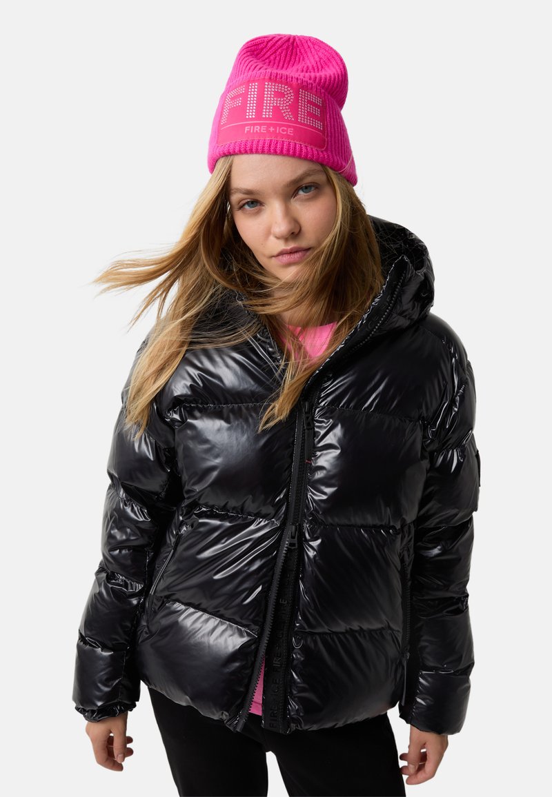 Ung kvinde med langt blond hår iført en skinnende sort pufferjakke og en lys pink hue med teksten "FIRE", der står med fronten mod kameraet.