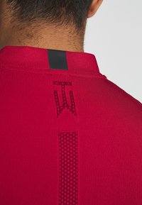 Röd idrottströja med ett strukturerat stickat mönster, utrustad med en svart accentremsa vid kragen och subtilt logodetalj på baksidan.