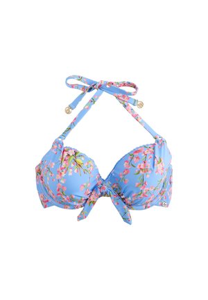 Donna che indossa un bikini blu a fantasia floreale, in piedi accanto a un muro di pietra con fiori rosa sullo sfondo.