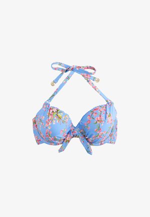 Frau trägt einen blauen Bikini mit Blumenmuster und steht neben einer Steinmauer mit pinkfarbenen Blumen im Hintergrund.