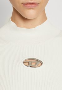 Hvit ribbestrikket topp med høy hals, som har en sirkulær utskjæring akkompanjert av en metallisk logo detalj i midten.