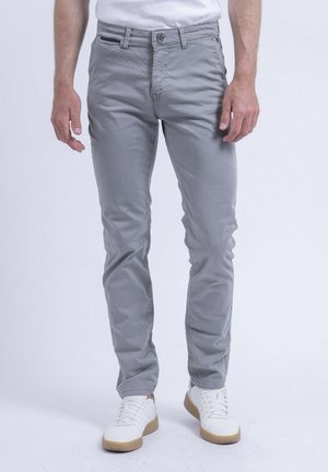 Homme portant un pantalon slim gris et des baskets blanches, debout devant un fond clair uni.