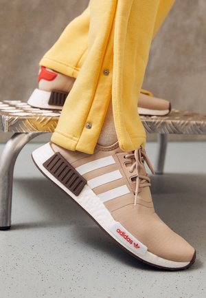 Beżowe buty sportowe Adidas z białymi paskami i czerwonym logo, noszone z żółtymi spodniami, jedna stopa oparta na metalowej ławce.