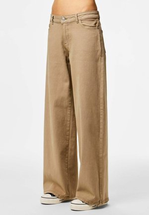 Béžové široké nohavice nosené osobou s viditeľným pásom, doplnené čierno-bielymi nízkoprofilovými teniskami, na jednoduchom pozadí.