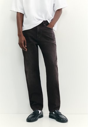 Person wearing black straight-leg jeans, white t-shirt tucked in front, white socks, and black loafers, standing against a white wall.