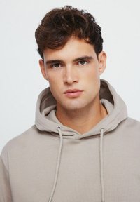 Beige Hoodie mit Kordelzug, der eine weiche Textur und eine lockere Passform aufweist. Das Design umfasst eine Kapuze und einen einfachen, minimalistischen Stil.