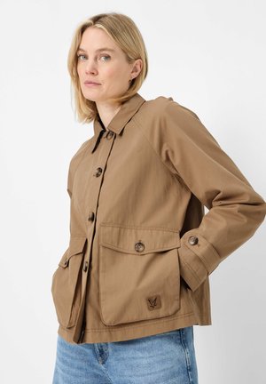 Frau mit blondem Haar, die eine beigefarbene Jacke mit großen aufgesetzten Taschen und blauen Jeans trägt, steht vor weißem Hintergrund.