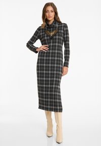 Langärmliges karriertes Kleid in Schwarz und Grau, mit einem Kragen und einem goldenen Adler-Akzent am Kragen. Kombiniert mit beigefarbenen kniehohen Stiefeln.