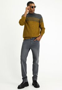 Senf- und grauer Strickpullover mit gestreiftem Design, kombiniert mit grauen Jeans und schwarzen Schnürschuhen. Accessoires mit runden Sonnenbrillen.