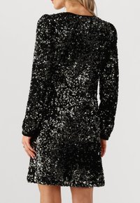 Robe noire à sequins avec manches longues, col rond, et ourlet légèrement évasé. Présente des sequins argentés réfléchissants sur l'ensemble.