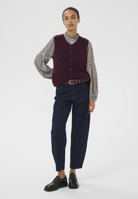 Bordeaux gebreide vest over een zwart-witte geruite blouse, gecombineerd met donkerblauwe high-waisted jeans en zwarte instappers.