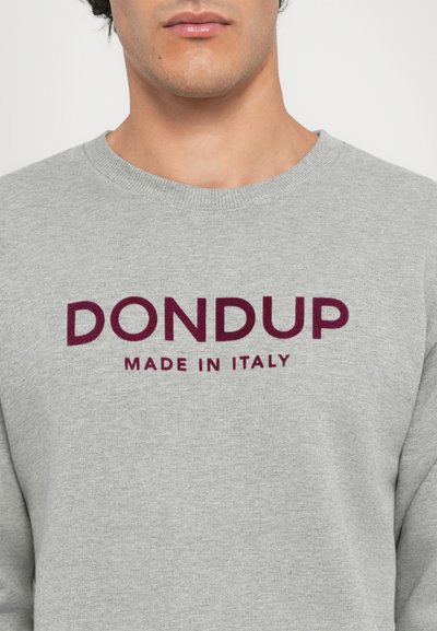 Vīrietis valkā gaiši pelēku sporta krekls ar tumši sarkano tekstu "DONDUP MADE IN ITALY", kas centrēts uz krūtīm.