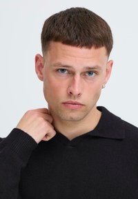 Črn pleten pulover z V-izrezom, ki ima teksturirano površino in rebraste manšete. Model ima kratke lase in uhane.