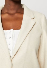 Crème-kleurige wolmix blazer met lapels en een zachte textuur, gecombineerd met een witte, geribbelde top met zichtbare knopen.