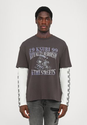 Dunkelgraues Kurzarm-T-Shirt mit grafischem Druck, das Text und eine Illustration zeigt. Wird über einem weißen Langarmshirt mit gemusterten Akzenten getragen.