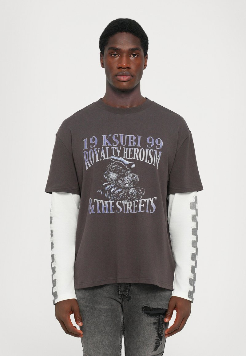 Donkergrijze T-shirt met korte mouwen, bedrukt met tekst en een illustratie. Draagt over een witte T-shirt met lange mouwen met patroonaccenten.