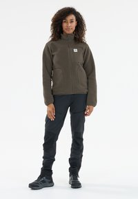 Brown fleece jacket with zip front, two side pockets, and a chest pocket. Black outdoor pants and sturdy black hiking boots.