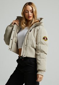 Beige cropped pufferjack met een met bont gevoerde capuchon, drukknoopsluiting, ritszakken en een Canada-patch op de mouw. Textuurstof.