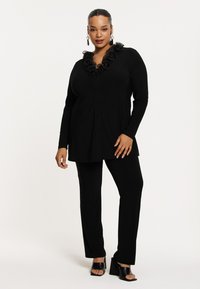 Schwarze Bluse mit Rüschensaum, kombiniert mit passenden schwarzen Hosen und klobigen schwarzen Sandalen mit Absatz vor einem neutralen Hintergrund.