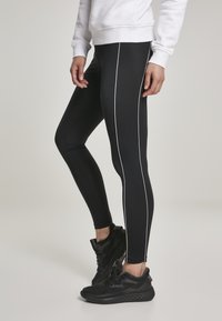 Fekete leggings fehér oldalsávokkal, rugalmas anyagból készült. Fekete sportcipőkkel párosítva. Egyszerű és testhezálló dizájn.
