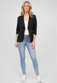Svart åpen front blazer med trekvartlange ermer, kombinert med lyseblå høy midje skinny jeans og hvite sneakers. Glatt stofftekstur.