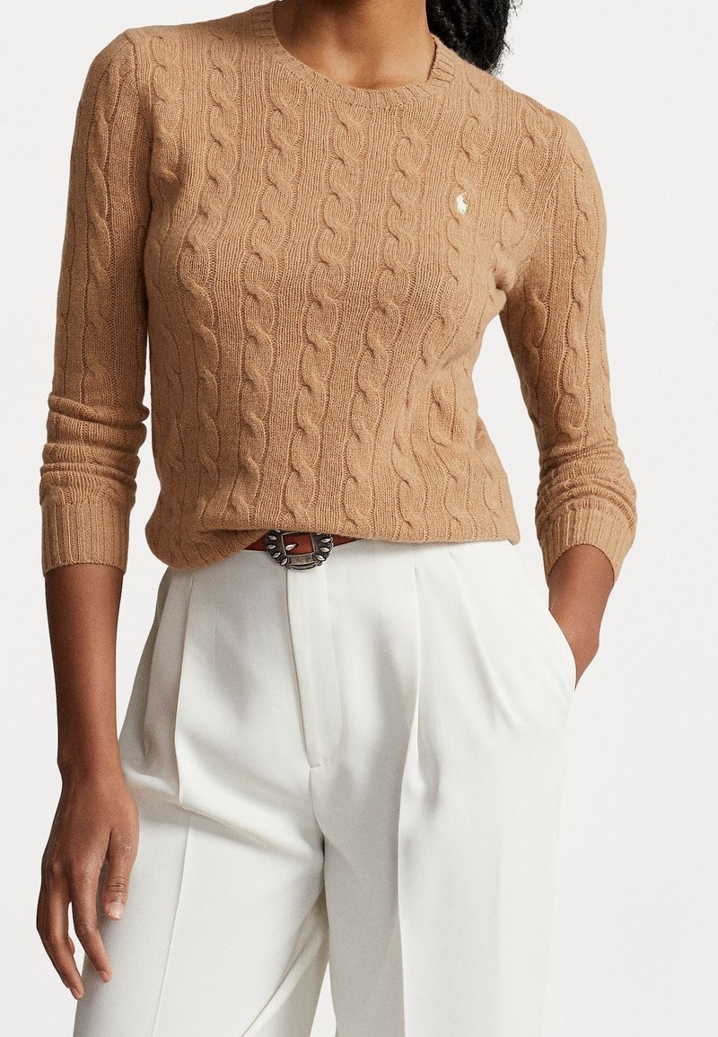 Femme portant un pull en maille torsadée beige avec un petit logo, rentrée dans un pantalon blanc plissé taille haute avec une boucle de ceinture décorative.