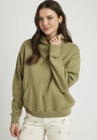 Haut à capuche vert olive en tissu doux, doté d'une poche avant, de poignets et d'un ourlet côtelés. Le modèle a les cheveux longs et porte un pantalon à motifs clairs.