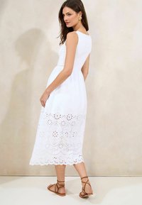 Robes sans manches blanches avec un corsage ajusté et une jupe évasée. Détails en dentelle sur le bord. Le modèle porte des sandales à brides marron.