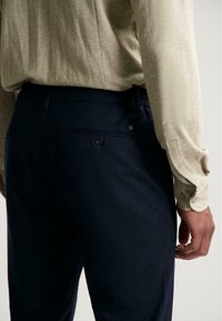 Man in een beige overhemd met lange mouwen, gestoken in donkerblauwe broek met knoopzakken aan de achterkant, staand tegen een effen achtergrond.
