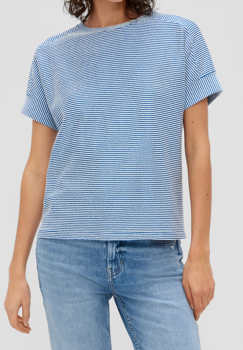 Femme portant une chemise à manches courtes rayée bleu et blanc et un jean bleu clair, debout, les bras détendus le long du corps, sur un fond uni.