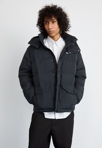 Schwarze Steppjacke mit Kapuze, mit einem Reißverschluss vorne, zwei großen Fronttaschen und elastischen Bündchen. Getragen über einem weiß gestreiften Hemd.