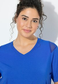 Mladá žena s kudrnatými tmavými vlasy svázanými do drdolu, která nosí malé kruhové náušnice a modrý top s výstřihem do V, jemně se usmívá na jednobílém pozadí.