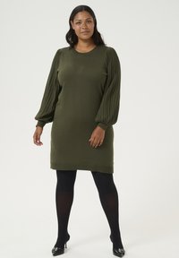 Robe pull vert olive avec des manches à côtes plissées et un col rond. Associée à des collants opaques noirs et des escarpins noirs à bouts pointus.