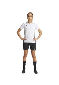 Maglietta sportiva bianca con accenti grigi, pantaloni corti neri e calze nere al ginocchio. Indossata con scarpe da calcio nere. Posizionato in avanti con le mani sui fianchi.