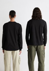 Zwei Personen stehen mit dem Rücken zur Kamera und tragen schwarze Langarmshirts; eine trägt beige Cargo-Hosen, die andere olivgrüne Cargo-Hosen.