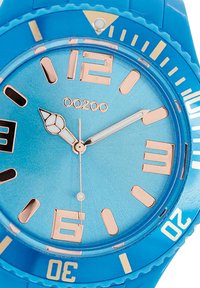 Montre en silicone bleue avec un cadran bleu métallisé, des index et aiguilles en or rose, et une lunette texturée. Présente un affichage de la date à 6 heures.