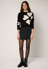 Jersey negro con patrones de corazones blancos, combinado con una minifalda oscura que incluye cinturón y hebilla. La modelo lleva botines negros y medias.