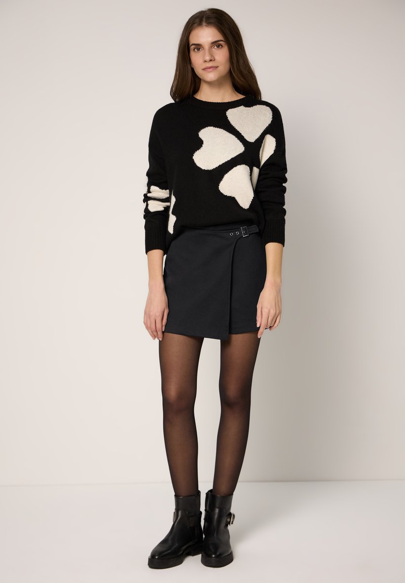 Jersey negro con patrones de corazones blancos, combinado con una minifalda oscura que incluye cinturón y hebilla. La modelo lleva botines negros y medias.