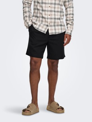 Homme portant une chemise à carreaux beige, un short noir et des sandales à enfiler couleur sable avec des détails de boucle, debout sur un fond uni.