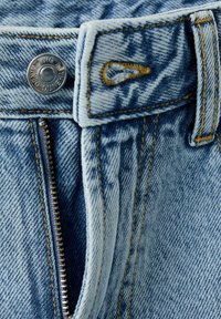 Gros plan d'un jean en denim bleu partiellement déboutonné montrant le bouton, la boutonnière, les coutures et les passants de ceinture.