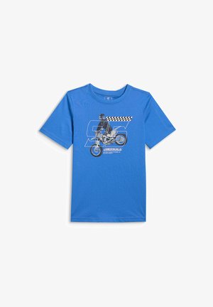 Chłopiec o blond włosach, ubrany w niebieską koszulkę z grafiką motocykla, czarne spodnie z niebieskimi paskami oraz czarno-białe trampki, stojący na szarym tle.