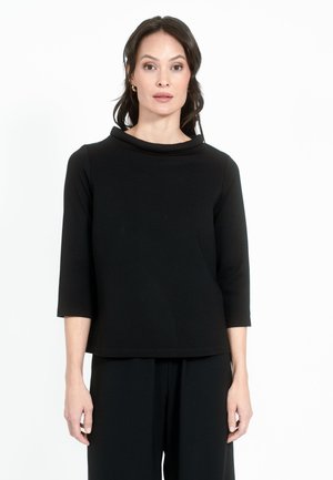 Maglietta nera con scollo a cowl e maniche a tre quarti, realizzata in tessuto morbido con una texture liscia e uniforme. Abbinata a pantaloni neri dritti.