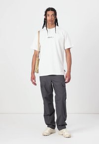 Camiseta blanca de algodón con logo negro, combinada con pantalones cargo grises con bolsillos laterales. Conjunto casual complementado con un bolso y zapatos beige.