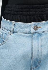 Κοντινή λήψη της μέσης ενός ανοιχτόχρωμου γαλάζιου τζιν με ασημένιο κουμπί με λογότυπο, φορεμένα πάνω από μαύρο ελαστικό παντελόνι.