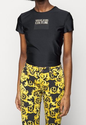 Personne portant un t-shirt noir cintré Versace Jeans Couture et un pantalon noir avec un motif baroque jaune orné.