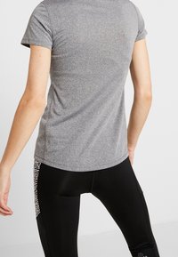 Camiseta atlética gris de manga corta con cuello redondo. Tela suave y transpirable. Combinada con leggings negros con un patrón decorativo en el lateral.